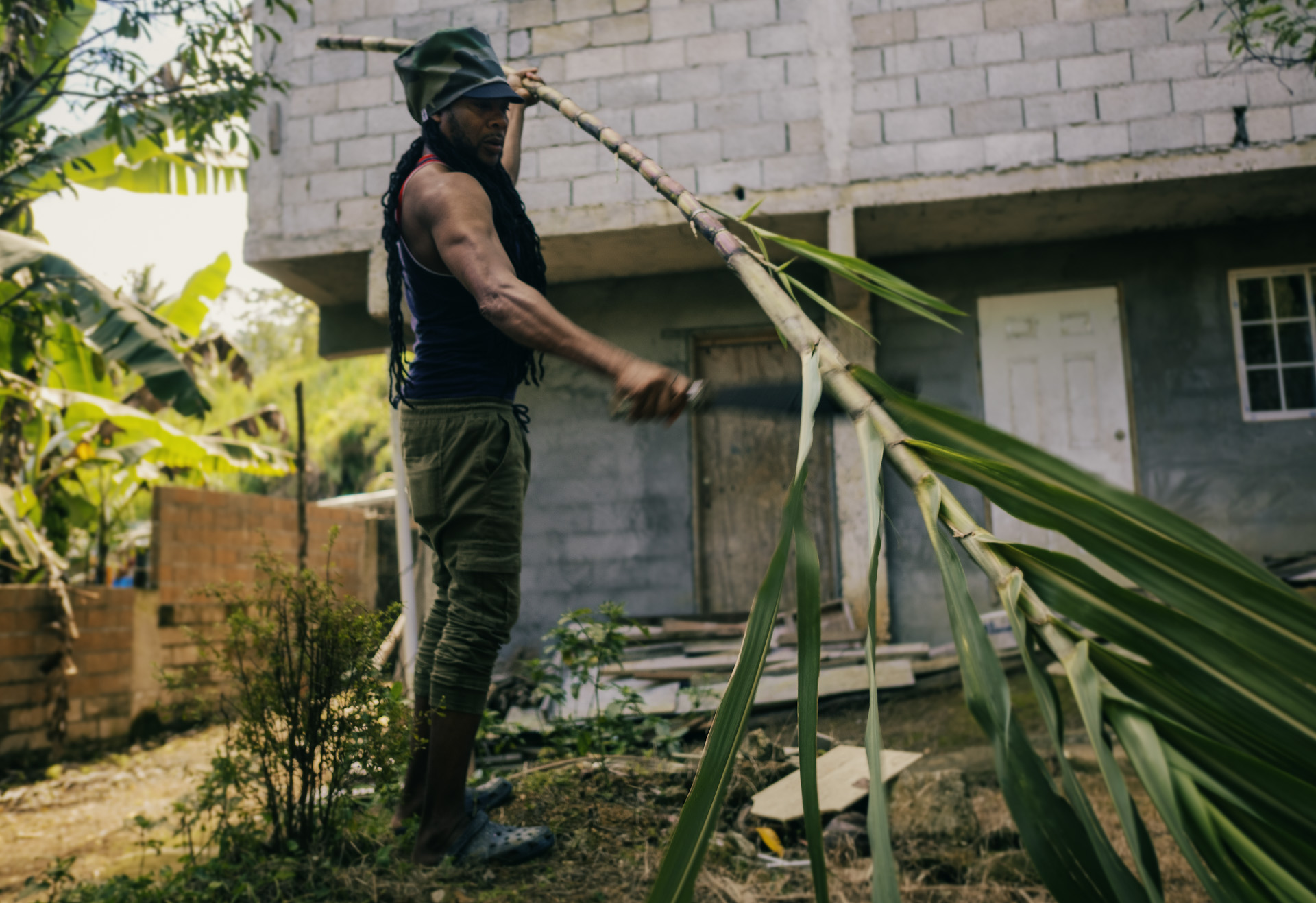 Patrick chopping the sugar cane in the backyard with his camo army dreadlocks cap