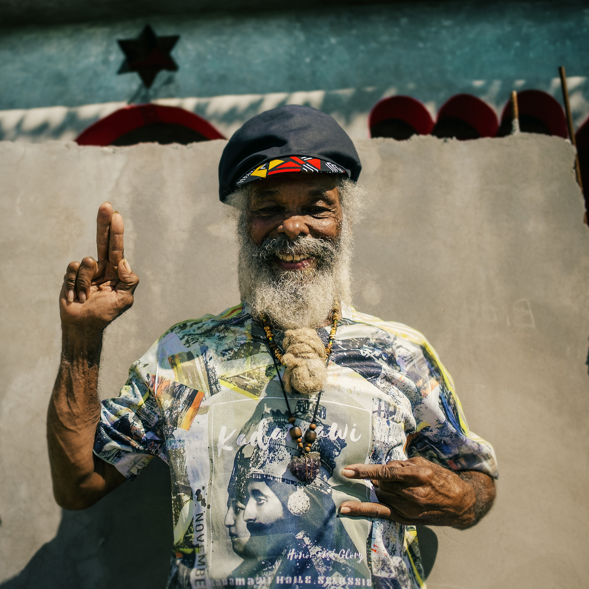 Noel Myton posing in front of his house and wearing a black dreadlocks hat with different colors in the visor