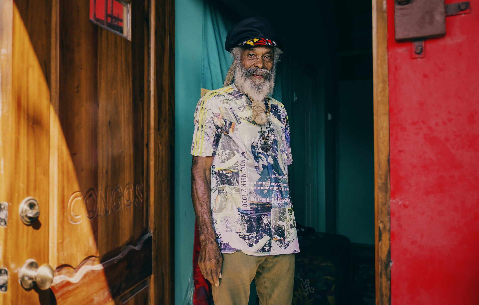 Noel Myton posing in front of Congos house and wearing a black dreadlocks hat with different colors in the visor