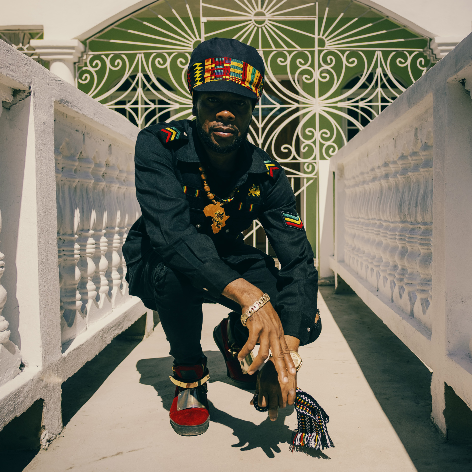 Proud Jamaican in front of a nice house wearing traditional and militant clothes striped by Ethiopian colors and a Dreadlocks Nation hat