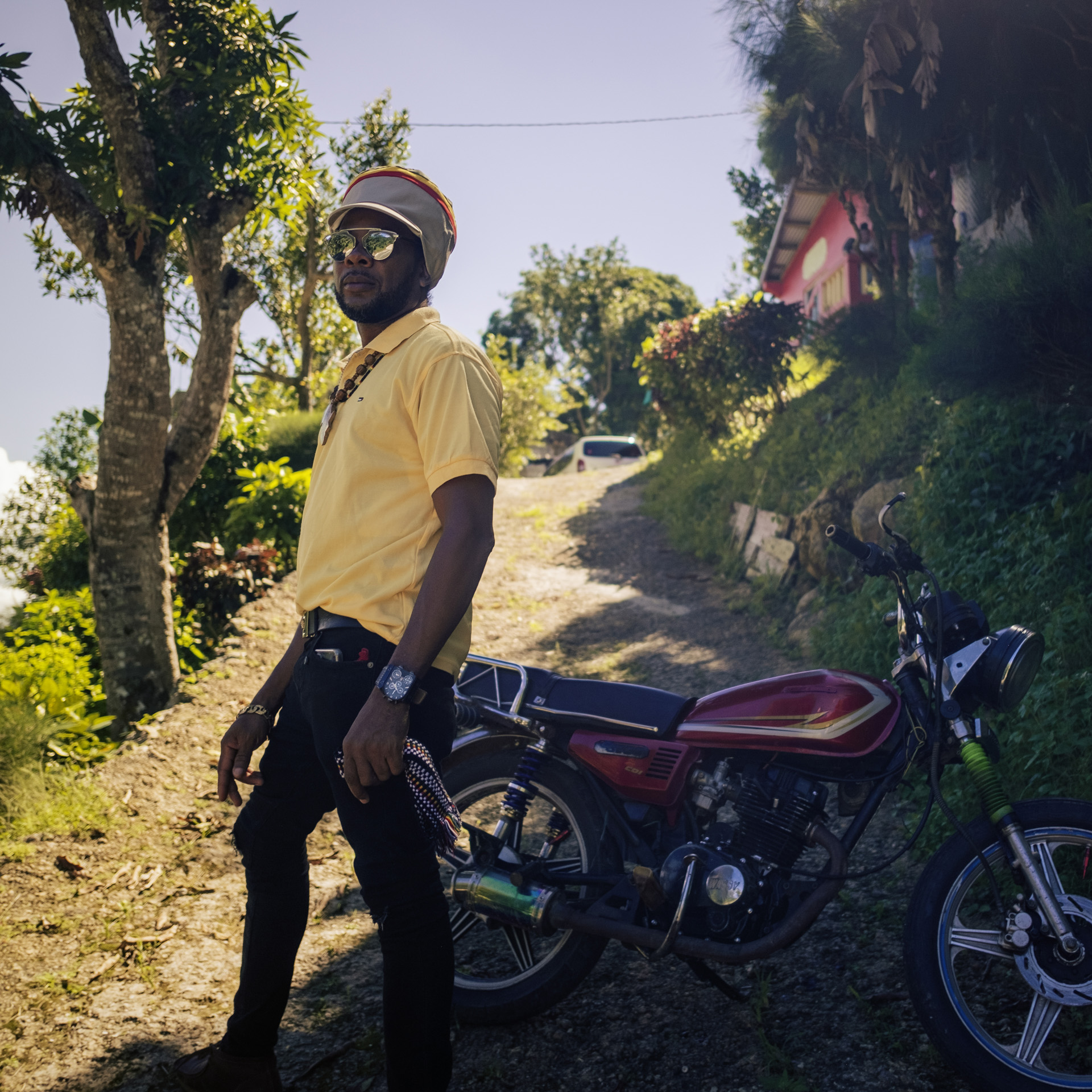 Patrique dans les rue de Trelawny devant une moto avec une casquette à Dreadlocks beige